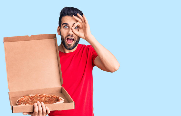 Молодой красивый мужчина с бородой держит доставку картона с итальянской пиццей улыбаясь счастливо делает хорошо знак с рукой на глазу глядя сквозь пальцы  - Фото, изображение