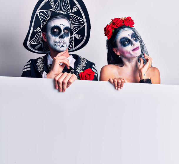 Jong stel draagt Mexicaanse dag van het dode kostuum met lege spandoek met de hand op kin denkend aan vragen, pensieve uitdrukking. glimlachend met een bedachtzaam gezicht. twijfelconcept.  - Foto, afbeelding