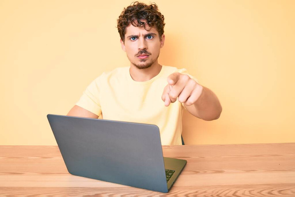 Junger kaukasischer Mann mit lockigem Haar im Büro, Laptop zeigt mit dem Finger in die Kamera und zu Ihnen, selbstbewusste Geste, die ernst wirkt  - Foto, Bild