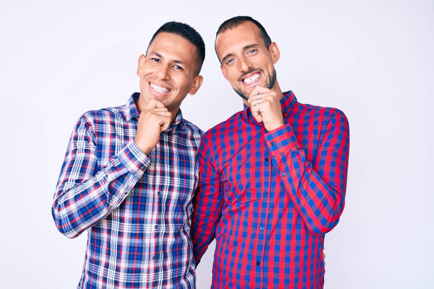 Молодая пара геев, одетых в повседневную одежду, уверенно смотрит в камеру, улыбаясь со скрещенными руками и поднятой на подбородок рукой. Позитивное мышление.  - Фото, изображение