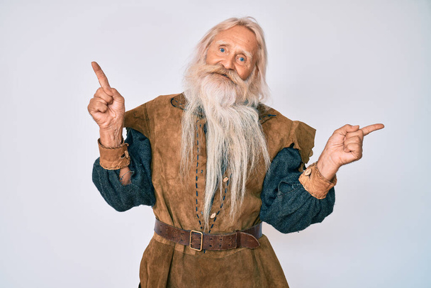Vieil homme âgé aux cheveux gris et à la barbe longue portant un costume traditionnel viking souriant confiant pointant avec les doigts vers différentes directions. espace de copie pour la publicité  - Photo, image