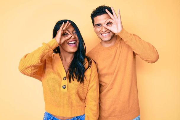 Beautiful latin young couple wearing casual clothes together doing ok gesture with hand smiling, eye looking through fingers with happy face.  - Photo, Image