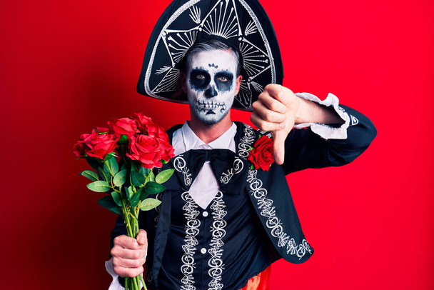 Молодой человек, одетый в мексиканский день мертвого костюма, держит розы с сердитым лицом, негативный знак, показывающий неприязнь с опущенными пальцами, концепция отказа  - Фото, изображение