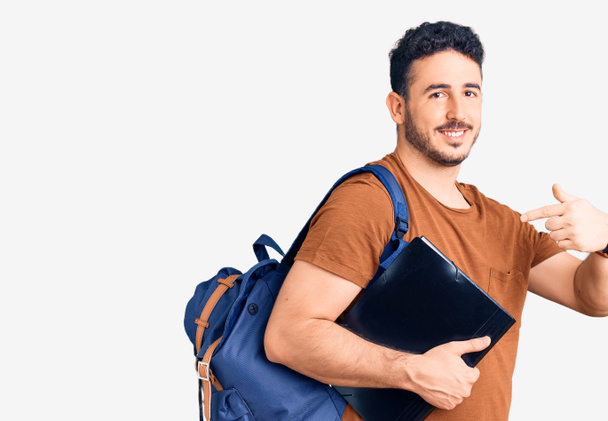 Joven hombre hispano usando mochila de estudiante sosteniendo aglutinante apuntando con el dedo a uno mismo sonriendo feliz y orgulloso  - Foto, imagen