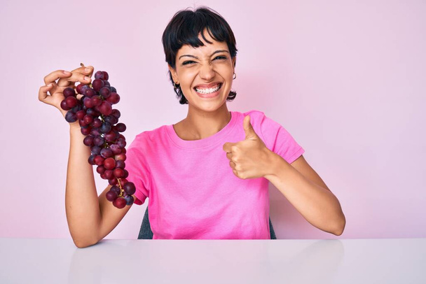Hermosa morena mujer sosteniendo rama de uvas frescas sonriendo feliz y positivo, pulgar hacia arriba haciendo excelente y signo de aprobación  - Foto, Imagen