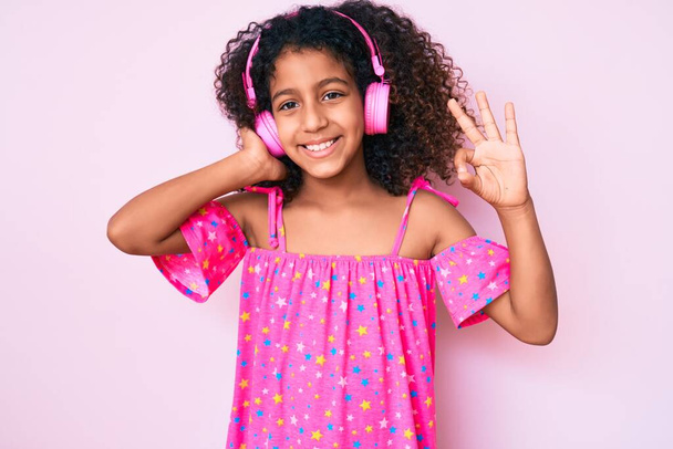 Afrikkalainen amerikkalainen lapsi, jolla on kiharat hiukset kuunnellen musiikkia kuulokkeilla, jotka tekevät ok-merkin sormilla, hymyilevät ystävällisiä eleitä erinomainen symboli  - Valokuva, kuva