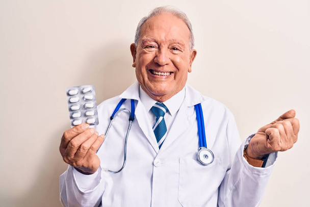 Starší šedovlasý lékař muž ve stetoskopu drží pilulky nad bílým pozadím křičí hrdě, slaví vítězství a úspěch velmi vzrušený se zvednutou paží - Fotografie, Obrázek