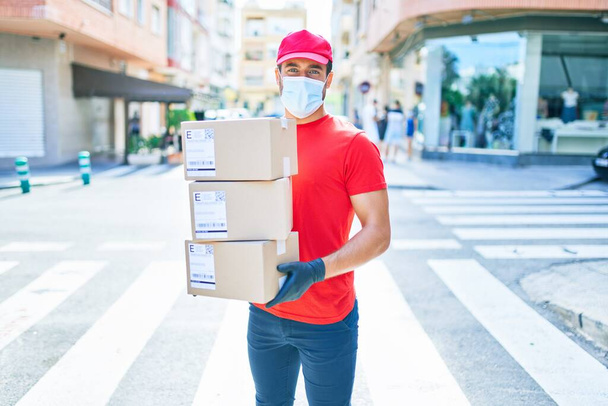 Νεαρός διανομέας φορώντας στολή και coronavirus προστασία ιατρική μάσκα κρατώντας πακέτα χαρτόνι στην πόλη δρόμο. - Φωτογραφία, εικόνα