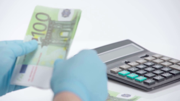 白い背景に計算機の近くにユーロ紙幣を数えるラテックス手袋の男 - 映像、動画