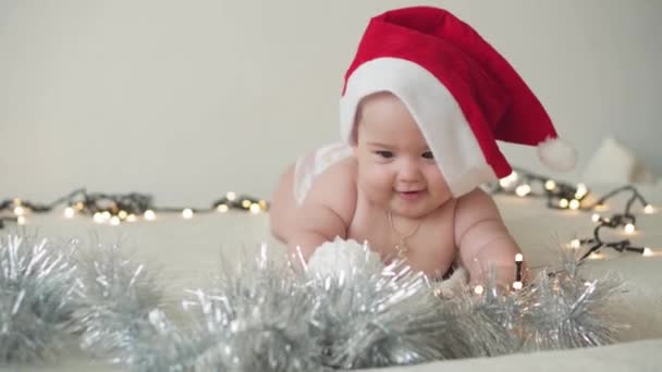 merry christmas christmas and happy new year, infants, childhood, holidays concept - close-up naked 6 month old newborn baby in santa claus hat on his tummy crawls with decorations on christmas tree. - Filmagem, Vídeo