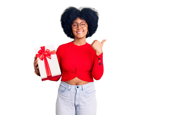 Giovane ragazza afroamericana che tiene in mano il regalo indicando la schiena dietro con mano e pollice in su, sorridendo fiducioso  - Foto, immagini