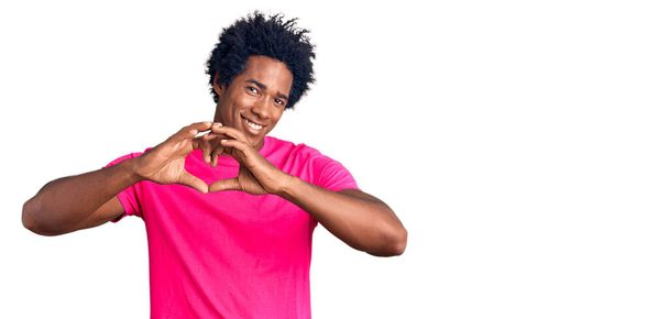 Knappe Afro-Amerikaanse man met afro haar in casual roze tshirt glimlachend in liefde met hart symbool en vorm met handen. romantisch concept.  - Foto, afbeelding