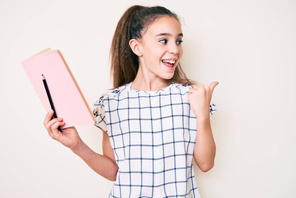 Cute hispanic child girl holding book and pencil pointing thumb up to the side smiling happy with open mouth  - Photo, Image