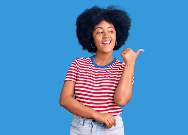 Νεαρή Αφροαμερικανή που φοράει καθημερινά ρούχα χαμογελώντας με χαρούμενο πρόσωπο και δείχνοντας στο πλάι με τον αντίχειρα προς τα πάνω.  - Φωτογραφία, εικόνα
