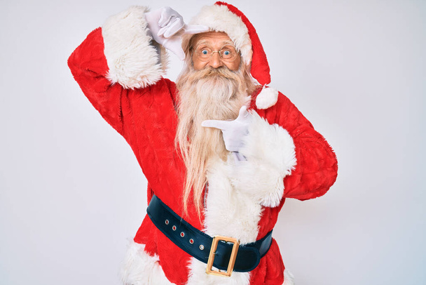 Vieil homme âgé aux cheveux gris et à la barbe longue portant un costume de Père Noël avec des bretelles souriantes faisant cadre avec les mains et les doigts avec un visage heureux. concept de créativité et photographie.  - Photo, image