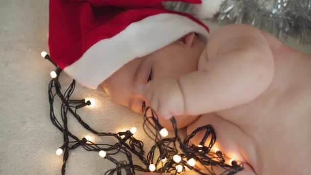 merry christmas christmas and happy new year, infants, childhood, holidays concept - close-up naked 6 month old newborn baby in santa claus hat on his tummy crawls with decorations on christmas tree. - Video