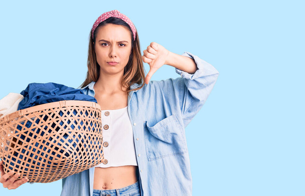 Young beautiful girl holding laundry basket with angry face, negative sign showing dislike with thumbs down, rejection concept  - 写真・画像