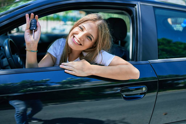 Νεαρή όμορφη ξανθιά γυναίκα χαμογελά ευτυχισμένη κάθεται στο αυτοκίνητο και δείχνει το κλειδί. - Φωτογραφία, εικόνα