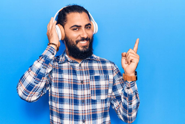 Молодий арабський чоловік слухає музику за допомогою навушників, усміхаючись, щасливо вказуючи рукою і пальцем на бік
  - Фото, зображення