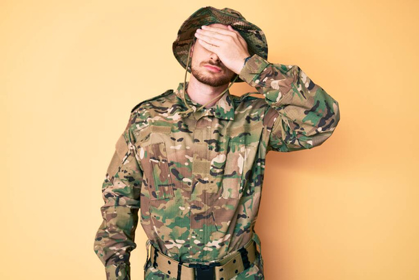 Young caucasian man wearing camouflage army uniform covering eyes with hand, looking serious and sad. sightless, hiding and rejection concept  - Zdjęcie, obraz