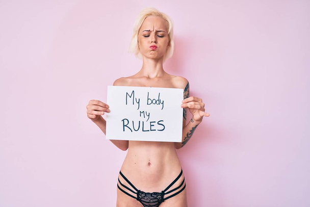 Young blonde woman with tattoo standing shirtless holding banner with my body my rules message making fish face with mouth and squinting eyes, crazy and comical.  - Fotografie, Obrázek