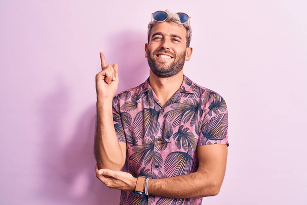 Bello biondo uomo in vacanza indossa camicia casual e occhiali da sole su sfondo rosa con un grande sorriso sul viso, puntando con mano e dito verso il lato guardando la fotocamera. - Foto, immagini