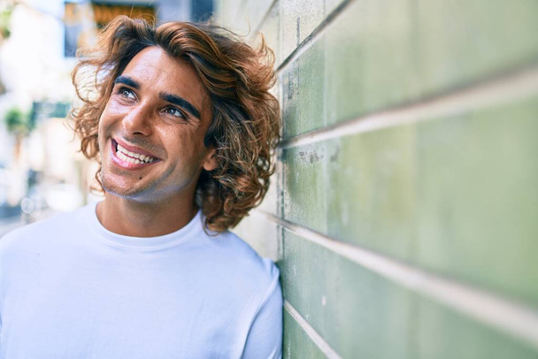 Молодой красивый латиноамериканец, счастливо улыбающийся, прислонившись к стене на улице города - Фото, изображение