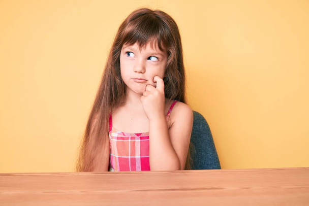 Petite fille enfant caucasienne aux cheveux longs portant des vêtements décontractés assis sur la table pensant concentré sur le doute avec le doigt sur le menton et regardant vers le haut se demandant  - Photo, image