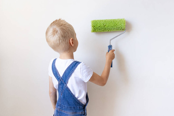 мальчик-строитель красит белые стены роликом в квартире, ребенок делает ремонт дома, вид сзади, место для текста - Фото, изображение