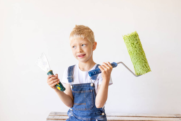 a blond child stands on a construction ladder in an apartment with white walls and a roller in his hands and looks into the distance, a place for text, the concept of repair - Photo, image
