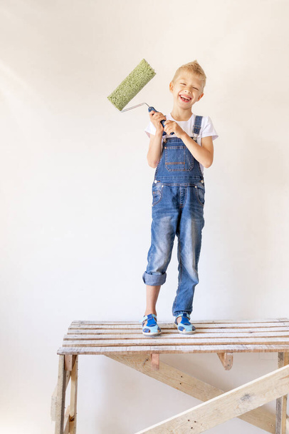 a blond child stands on a construction ladder in an apartment with white walls and a roller in his hands, a place for text, the concept of repair - Фото, изображение