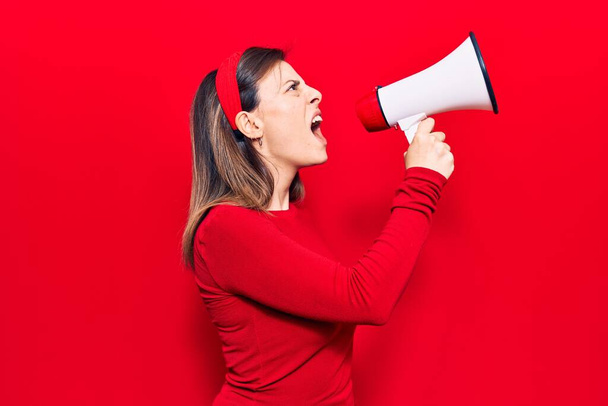 Νεαρή όμορφη γυναίκα που φοράει ντάιαντεμ με θυμωμένη έκφραση. Φωνάζοντας δυνατά χρησιμοποιώντας μεγάφωνο που στέκεται πάνω από απομονωμένο κόκκινο φόντο - Φωτογραφία, εικόνα