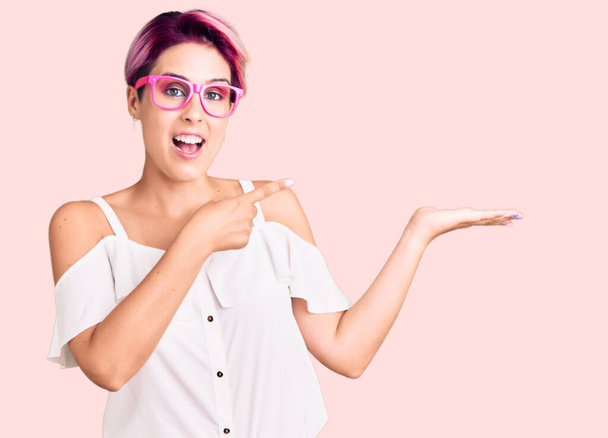 Jeune belle femme aux cheveux roses portant des vêtements décontractés et des lunettes étonnées et souriantes à la caméra tout en présentant avec la main et pointant du doigt.  - Photo, image