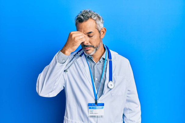 Keski-ikäinen harmaa tukka mies yllään lääkäri yhtenäinen ja stetoskooppi väsynyt hankausta nenä ja silmät tunne väsymystä ja päänsärkyä. stressi ja turhautuminen käsite.  - Valokuva, kuva