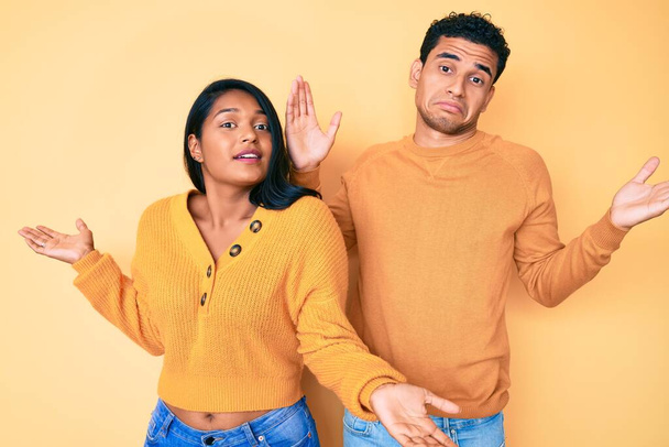 Beautiful latin young couple wearing casual clothes together clueless and confused expression with arms and hands raised. doubt concept.  - Photo, Image