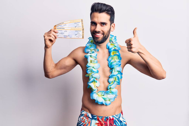 Young handsome man with beard wearing swimwear and hawaiian lei holding boarding pass smiling happy and positive, thumb up doing excellent and approval sign  - Foto, Imagem