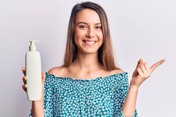 Young beautiful girl holding bottle of cream lotion smiling happy pointing with hand and finger to the side  - Фото, изображение