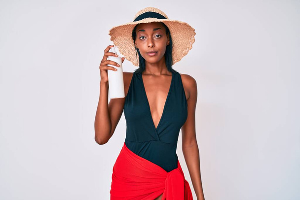 Genç Afrikalı Amerikalı kadın mayo giyiyor ve elinde güneş kremi tutuyor. Düşünme tavrı ve ayık yüz ifadesi. Kendine güveniyor gibi görünüyor.  - Fotoğraf, Görsel