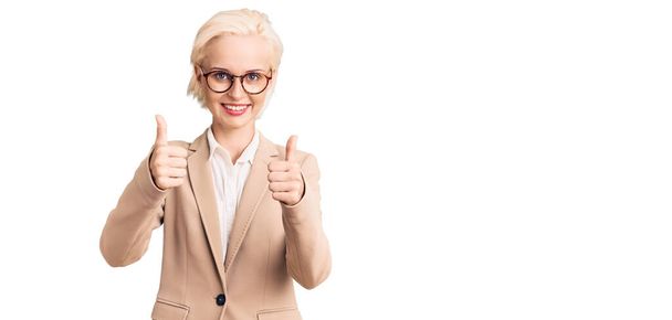 Jeune femme blonde portant des vêtements d'affaires et des lunettes signe de succès faire un geste positif avec la main, pouces levés souriant et heureux. expression joyeuse et geste gagnant.  - Photo, image