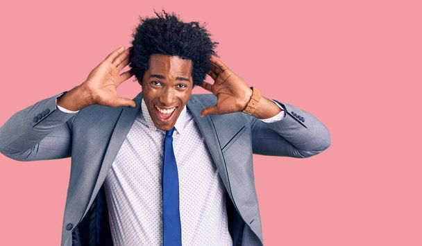 Красивый африканский американец с афроволосами в деловой куртке, улыбающийся, дергая за уши пальцами, смешной жест. проблема прослушивания  - Фото, изображение