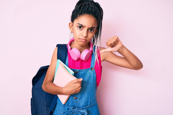 Mladý africký Američan dívka dítě s copánky drží studentský batoh a knihy s rozzlobený obličej, negativní znamení ukazuje odpor s palci dolů, odmítavý koncept  - Fotografie, Obrázek