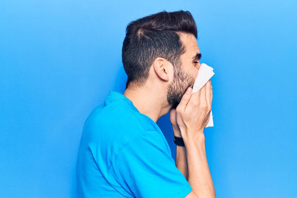 Enfermedad del joven hispano usando pañuelo de papel en la nariz. De pie sobre fondo azul aislado - Foto, imagen
