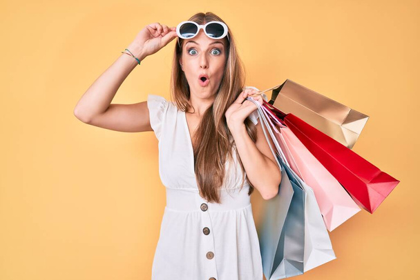 Young blonde girl holding shopping bags afraid and shocked with surprise and amazed expression, fear and excited face.  - Foto, imagen