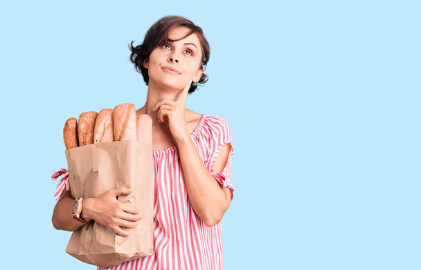 Gyönyörű fiatal nő rövid haj tartó papír táska kenyér komoly arc gondolkodás kérdés kézzel az állon, gondolkodó zavaros ötlet  - Fotó, kép