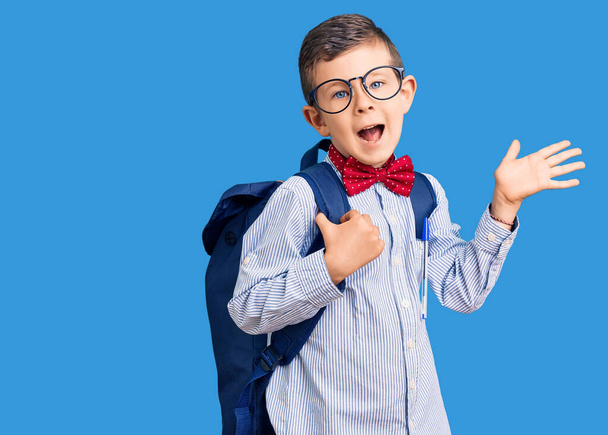 Cute blond kid wearing nerd bow tie and backpack celebrating victory with happy smile and winner expression with raised hands  - Foto, afbeelding