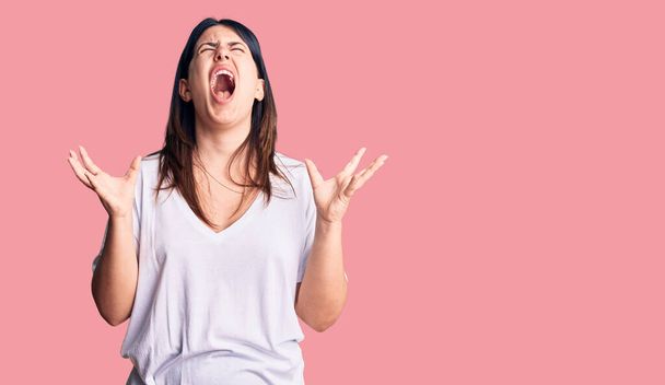 Young beautiful brunette woman wearing casual t-shirt crazy and mad shouting and yelling with aggressive expression and arms raised. frustration concept.  - Photo, Image