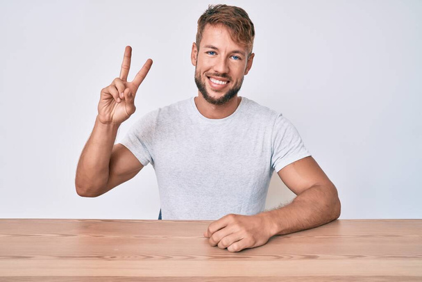 Young caucasian man wearing casual clothes sitting on the table smiling with happy face winking at the camera doing victory sign. number two.  - Photo, Image