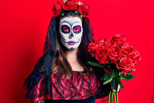 Young woman wearing day of the dead costume holding flowers thinking attitude and sober expression looking self confident  - 写真・画像