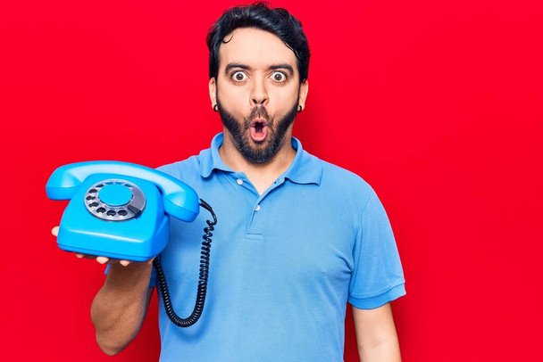 Jeune homme hispanique tenant un téléphone vintage effrayé et étonné avec la bouche ouverte pour la surprise, visage incrédule  - Photo, image