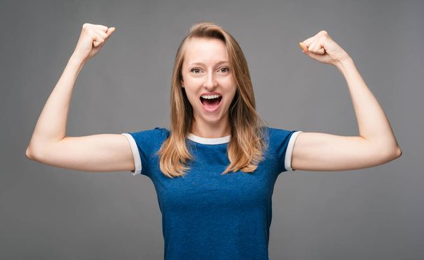 Successful Young female with blonde straight hair, dressed in blue t shirt, smiling happily, celebrating amazing news, making Winner gesture. Human emotions, facial expression concept. - Foto, imagen